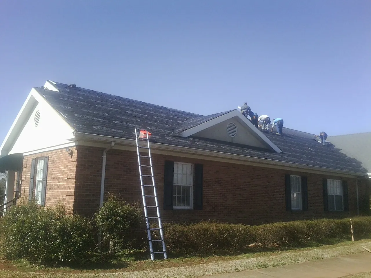 Crew tearing shingles off a brick ranch roof with a ladder set — Metal Roof Repair work by our crew in Rollins