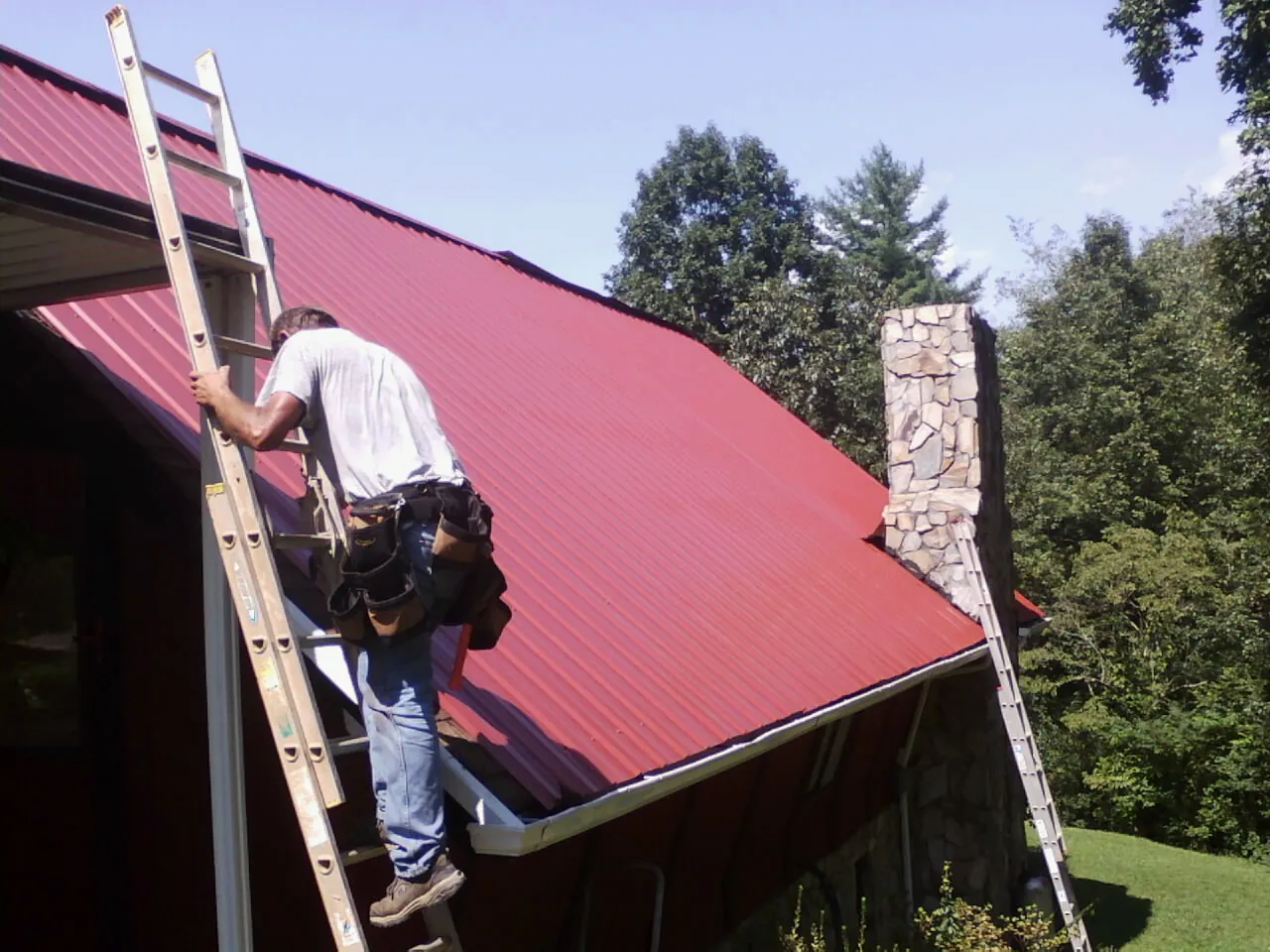 Roofer on a ladder beside a red metal roof and stone chimney — Soffit & Fascia Repair by Roofing Brothers in Rollins